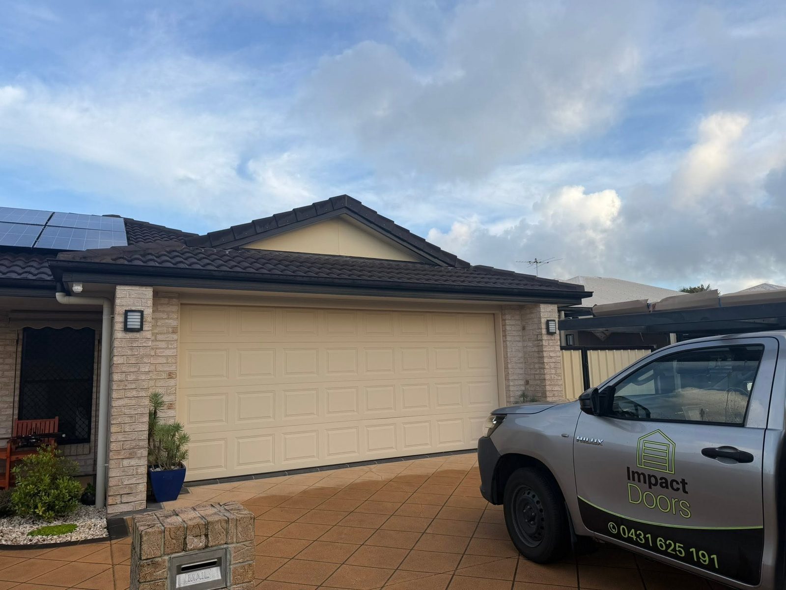 The branded Impact Doors ute parked in front of a cream panel-lift garage door on a brick home.