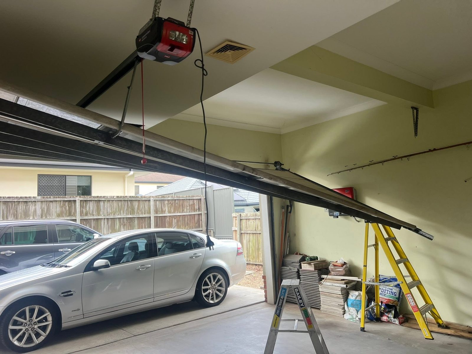 A garage door off its track from inside the garage, propped on a ladder above a parked car with the opener and rail overhead.