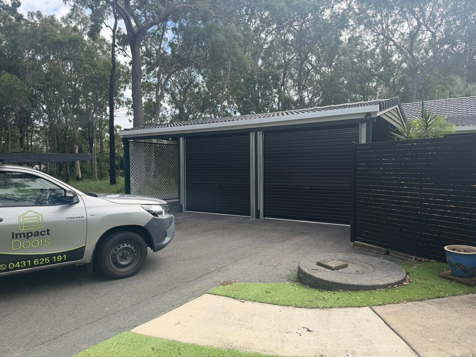 The branded Impact Doors ute in the driveway of a bushland property with two black roller doors on the garage.