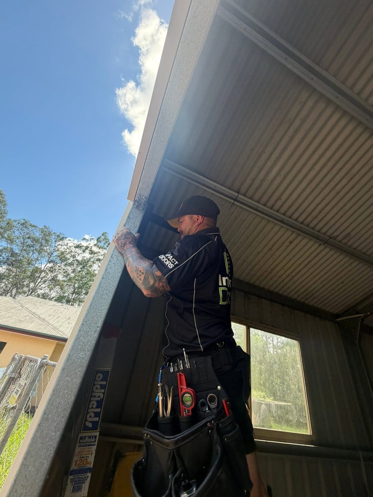 Jarred from Impact Doors working on a garage door track at the top of a ladder, wearing a branded shirt and tool belt.