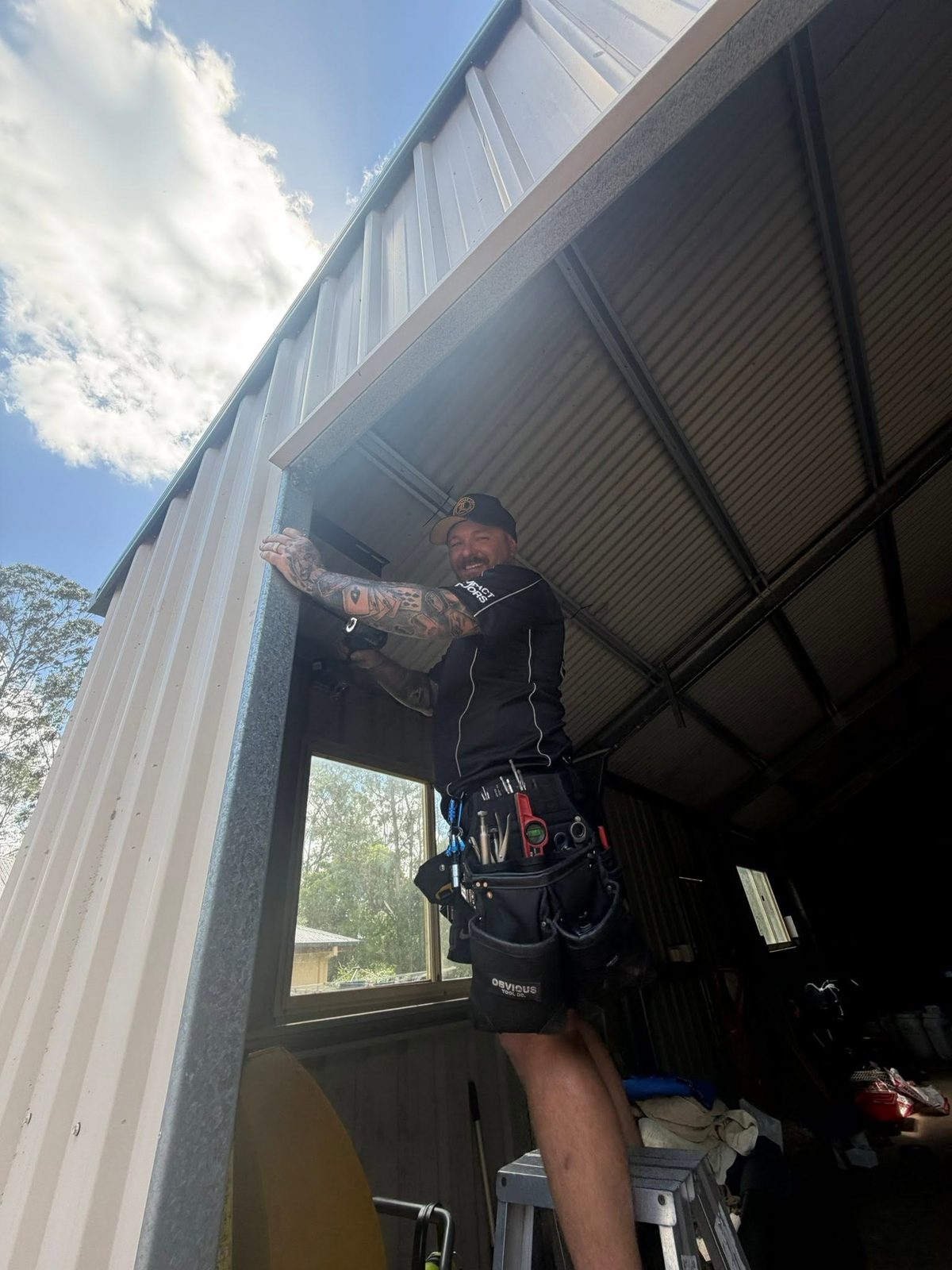 Jarred from Impact Doors standing on a ladder in a branded work shirt and tool belt, smiling at the camera while fitting a garage door opener.